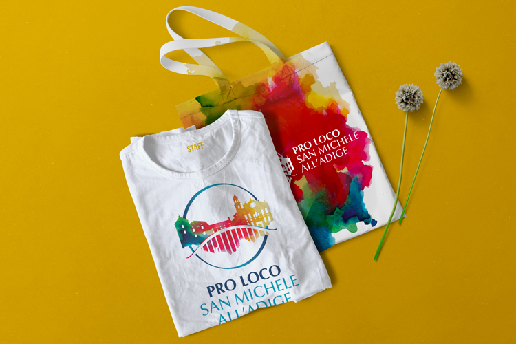 Realizzazione grafica Shopper e T-shirt Pro Loco San Michele all'Adige.La T-shirt bianca è caratterizzata dal logo, nella versione verticale, rappresentato dal profilo della chiesa del paese acquerellato: verde, blu, rosso, arancione e giallo. Il ponte sull'Adige sintetizzato e di colore bianco, si staglia sopra il profilo del paese. I due elementi chiave sono uniti da un cerchio dal colore sfumato blu. La Shopper bianca riprende la texture acquerellata del logo: verde, blu, rosso, arancione e giallo come sfondo e in primo piano è posizionato il logo, versione orizzontale, in negativo bianco.